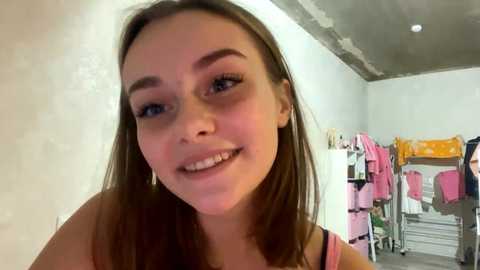 Video of a young woman with straight brown hair and light skin, smiling, wearing a pink top, standing in a small, cluttered room with a white ceiling and hanging clothes.