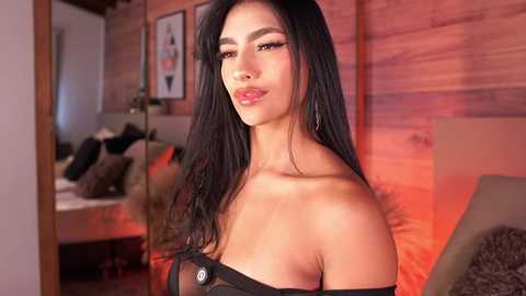 Video of an attractive, tan-skinned woman with long, black hair, wearing an off-shoulder black dress, posing confidently in a modern, warmly lit living room with wooden walls, cushions, and framed artwork in the background.