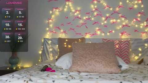 Video of a cozy bedroom with a white bed, adorned with pink and white pillows, and a pink blanket. The wall features a string of fairy lights and bird-shaped decorations, creating a romantic ambiance.