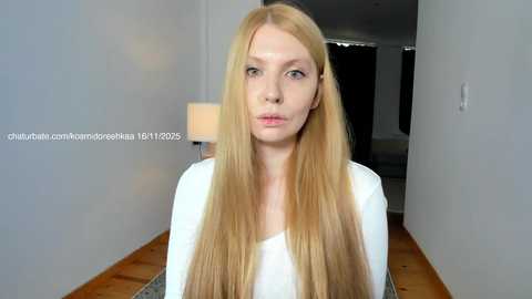 Video of a pale-skinned, blonde woman with long hair, wearing a white top, pouting in a modern, minimalist room with wooden floors and grey walls.