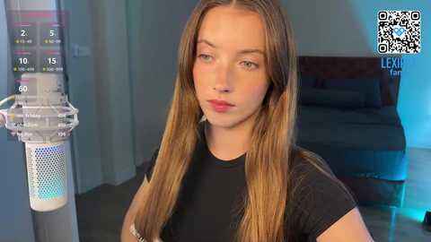 Video of a young, fair-skinned woman with long, straight, blonde hair, wearing a black t-shirt, standing in front of a white air purifier. Background shows a modern bedroom with a bed and blue lighting.
