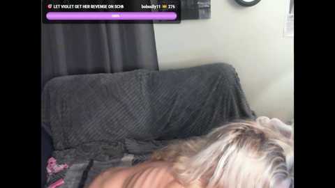 Video of a person with long, platinum blonde hair lying face down on a gray, textured blanket, with a purple and pink pillow visible. A black curtain and a wall-mounted clock are in the background.