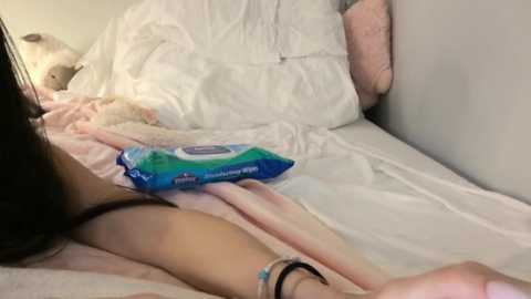 A video of a person lying on a bed with messy white sheets, wearing a black thong and a blue bag of chips, in a dimly lit room.