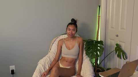 Video of a young Asian woman with medium build, small breasts, and dark hair in a bun, wearing a white sports bra and beige shorts, sitting on a white chair in a simple, dimly lit room with a green plant.