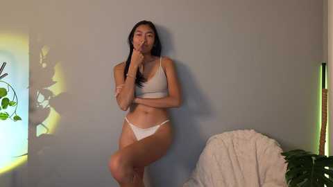 Video of a slim, light-skinned Asian woman with long black hair, wearing white lingerie, standing against a light gray wall, touching her lips.