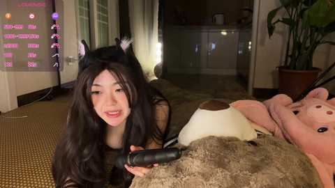 A video of a young Asian woman in a black cat costume, lying on a plush brown bear, holding a black vibrator, in a dimly lit living room with a TV and a plant.