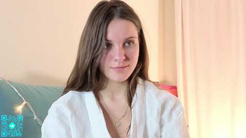Video of a young Caucasian woman with long brown hair, fair skin, and a slender physique, wearing a white robe, seated indoors with a soft, warm background.