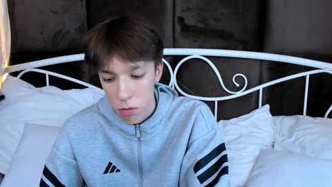 Video of a teenage boy with light skin, wearing a light gray Adidas jacket, lying on a bed with white sheets and a black metal headboard.