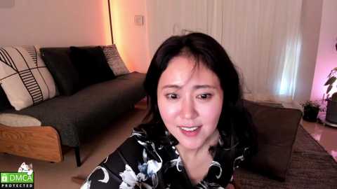 Video of an Asian woman with long black hair, wearing a black floral robe, sitting on a brown sofa in a dimly lit living room with beige walls and a potted plant.