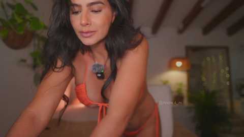 Video of a middle-aged woman with long, wavy black hair, wearing an orange bikini, adjusting the top in a dimly lit room with wooden beams and plants.