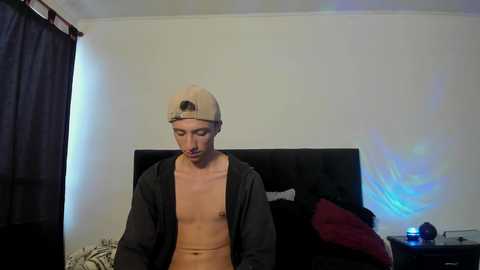 Video of a young, slim, shirtless man with light skin and short hair, wearing a backwards baseball cap, standing in a dimly lit bedroom with a black TV screen and a red blanket.