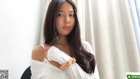 Video of an Asian woman with long black hair, light skin, and a slender figure, wearing a white off-shoulder top, adjusting her bra strap. She has a neutral expression.
