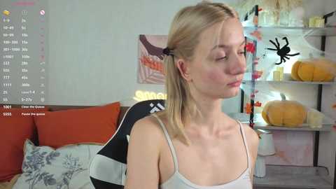 A video of a fair-skinned, slender young woman with blonde hair in pigtails, wearing a white spaghetti-strap top, seated in an orange chair in a cozy, cluttered room with Halloween decorations.