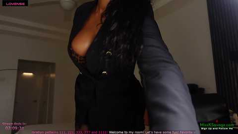 A video of a woman with long, wavy black hair, wearing a plunging black lace dress, posing in a dimly lit living room.