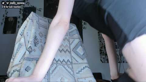 A video captures a light-skinned woman in a black top leaning over a patterned, light-colored couch. The background shows a cluttered, dimly lit room with abstract art.