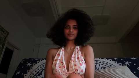 Video of a young woman with a voluminous afro and light brown skin, wearing a white halter top adorned with pink flowers. She sits on a blue and white patterned couch in a dimly lit room.