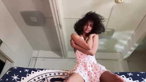 Video of a young Black woman with voluminous curly hair, wearing a white floral dress, sitting on a bed with a black and white patterned duvet, in a dimly lit room with white walls and ceiling.