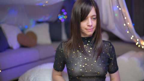 Media: Video of a fair-skinned woman with straight brown hair, wearing a sheer black top with gold speckles, smiling slightly in a cozy, dimly lit room with string lights and a heart-shaped pillow.