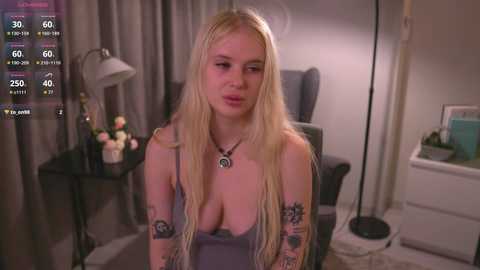 Media: Video of a blonde woman with long hair, wearing a low-cut grey top, seated in a dimly lit room with a lamp, flowers, and a black chair.