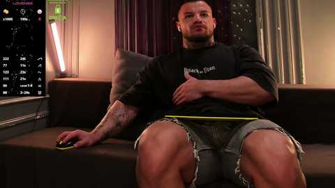 Video of a muscular, tattooed man in a black shirt and shorts, seated on a dark couch, holding a controller, in a dimly lit room with a calendar and cityscape background.