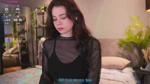 Video of a young woman with long dark hair, wearing a sheer black top, sitting on a bed in a cozy bedroom with a plant and a screen showing \"OF Think in my bio.\