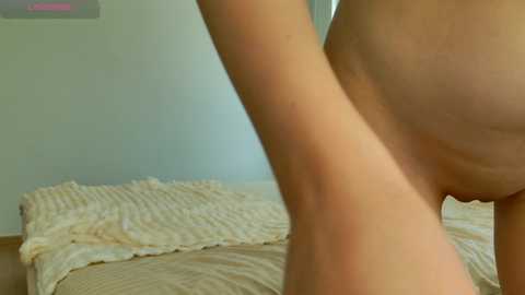 A close-up video of a nude person's torso and thighs, with light skin, standing in a sparsely decorated bedroom. The bed has a cream-colored blanket and beige walls.