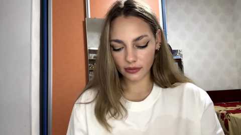 Video of a young Caucasian woman with long, straight, blonde hair, wearing a white t-shirt, standing in a brightly lit room with orange walls and a bed with red and gold bedding in the background.
