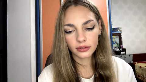 Video of a young woman with long, straight, light brown hair, wearing white clothing, with closed eyes, and black eyeliner. Background includes an orange wall and a colorful painting.