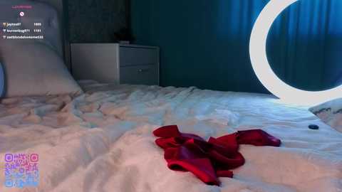 Video of a disheveled bed with white sheets, a red bra, and a phone, set against a teal wall with a circular ring light.