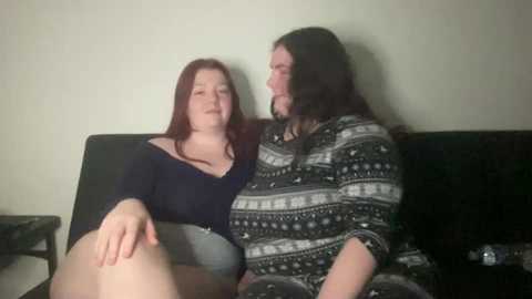 Video of a pale, red-haired woman in a dark top and gray shorts, sitting on a black couch next to a bearded man in a patterned shirt.