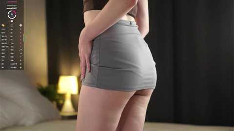 Video of a light-skinned woman in a tight, grey mini skirt, standing sideways, showing off her toned thighs and butt. She's in a modern bedroom with a bed, lamp, and dark curtains in the background.