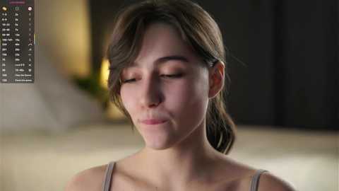 Video of a young woman with light skin, brown hair tied back, wearing a gray tank top, with closed eyes and a relaxed expression, in a softly lit, blurred room.