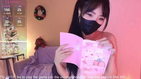 Media: A video captures an East Asian woman with long black hair, wearing a black mask and pink cardigan, holding a pink card with cartoonish food. The background features a festive room with a Christmas tree and a warm, cozy atmosphere.