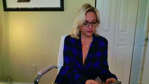 Video of a blonde woman in glasses, wearing a blue plaid shirt, seated at a desk, in a beige-walled office with framed art and white door in the background.