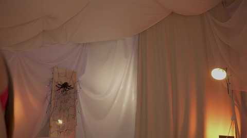 Video of a dimly lit, draped room with white and beige curtains, a small table, and a circular lamp casting a warm glow, creating a soft, romantic ambiance.