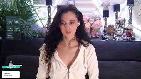 Video of a young woman with long dark hair, wearing a white zip-up hoodie, seated on a black sofa. Background includes a monitor displaying \"Training\" and \"Zachary,\" with toy figurines and skulls.