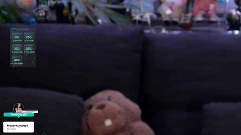 A video showing a dark gray couch with a plush brown bear on it. The couch displays a virtual reality interface with health stats. Background features a cluttered room with various objects.