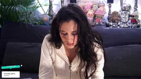 Video of a woman with long dark hair, wearing a white hoodie, sitting on a black couch in a room with plush toys and plants.