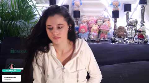 Video of a woman with long black hair wearing a white hoodie, seated in a modern living room with Star Wars figurines and plants in the background.