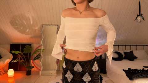 Video of a slender, light-skinned woman with short brown hair, wearing a white off-the-shoulder crop top and black patterned pants, standing in a cozy, dimly lit attic bedroom with wooden walls, a bed, and a potted plant.