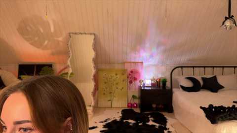 Video of a cozy, warmly lit bedroom with a white ceiling, a black cowhide rug, a bed with black pillows, and a mirror reflecting a person's face.