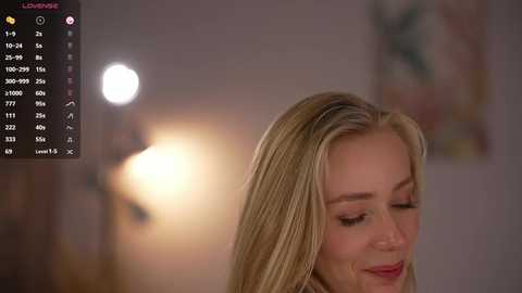 Video of a smiling blonde woman with long hair, wearing makeup, in a softly lit room with blurred, pastel-colored wall art in the background.