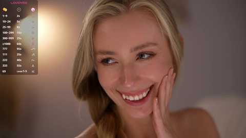 Video of a smiling blonde woman with light skin, wearing a white top, hand on her cheek, in a softly lit room.