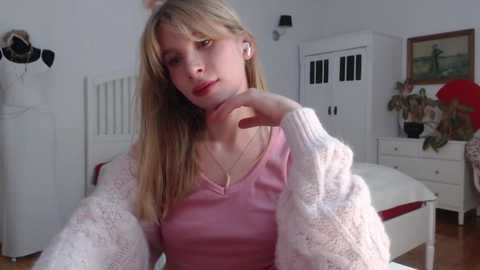 Video of a young, fair-skinned, blonde-haired woman in a pink sweater, sitting on a bed with white linens in a minimalist bedroom with white furniture.