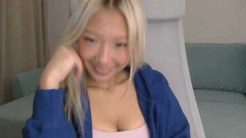 Video of a smiling East Asian woman with long blonde hair, wearing a blue cardigan over a pink top, seated on a light blue couch in a room with beige walls.