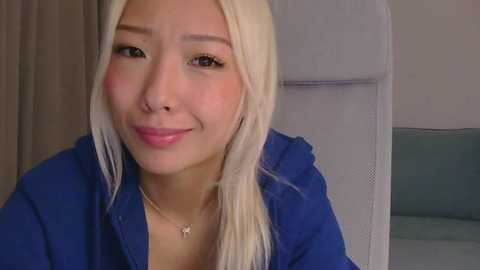 Video of a smiling Asian woman with long, straight platinum blonde hair, fair skin, and wearing a blue shirt, taken indoors, focusing on her face.