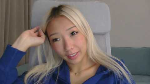 Video of an Asian woman with long, platinum blonde hair and light skin, wearing a blue shirt, smiling while touching her hair, in a modern, light-colored room with a grey chair.