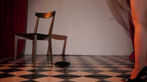 Video of a minimalist room with a black-and-white checkered floor, a single wooden chair, and a black garment discarded on the floor, against a white wall with red curtains.