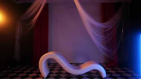 Video of a white, curvaceous, abstract sculpture on a black-and-white checkered floor. The background features sheer, flowing pink and red drapes against dark walls, with soft, warm lighting creating a dreamy atmosphere.
