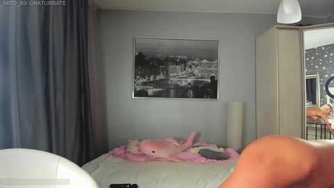 A video of a modern bedroom with a gray wall, a large framed black-and-white photo of a cityscape, a plush pink shark toy on a white bed, and a white lamp beside it.